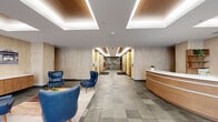 Located at 332 Dayton St in Hamilton, Ohio, this polished commercial lobby presents a sophisticated and welcoming environment thoughtfully designed for professional use. The single-floor space encompasses a generous footprint that allows for a well-organized layout with distinct functional zones flowing seamlessly from one to the next.

Upon entry through a floor-to-ceiling glass and steel vestibule, visitors are greeted by an abundance of natural light streaming in from the expansive glazed facade. The interior features large-format stone tile flooring laid in a subtle grid pattern, lending a sense of refinement throughout. Warm travertine-style wall cladding lines the perimeter, complemented by wood accents on the ceiling coffers and built-in millwork, creating a balanced blend of contemporary and classic materials.

A curved wood-and-white reception desk serves as the focal point, positioned to welcome guests efficiently. Adjacent to it, a comfortable seating area features mid-century-inspired blue leather armchairs arranged around a low coffee table, with local black-and-white photography displayed on the walls. A wall-mounted television adds a functional element near the entrance.

Toward the rear, a corridor lined with gold-toned elevator doors leads deeper into the building, branded with "Third + Dayton" signage. Recessed linear lighting throughout enhances the clean, professional ambiance of this well-appointed lobby.