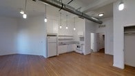 This single-floor loft space showcases a striking industrial aesthetic that blends raw architectural character with functional design. The most immediately notable features are the soaring ceilings with exposed cylindrical ductwork running overhead, lending an authentic urban loft feel throughout the open-concept interior.

The kitchen is positioned along one wall and is outfitted with white cabinetry, open shelving, a white refrigerator, and a white range with oven. A stainless steel sink sits beneath upper shelving, and the white appliances and cabinetry create a clean, bright contrast against the surrounding industrial elements. Above the kitchen area, a row of pendant Edison-style bulb fixtures hangs at varying lengths, providing both task lighting and a design focal point that reinforces the loft's industrial character.

The flooring throughout is wide-plank hardwood in a warm honey tone, adding natural warmth and texture that balances the otherwise utilitarian overhead elements. The walls are finished in crisp white, maximizing light reflection and enhancing the sense of openness. A hallway visible at the rear of the space leads to additional rooms, suggesting further functional areas beyond the main open floor. The overall ambiance is spacious, airy, and well-suited to a modern, urban lifestyle.