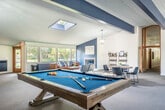 Located at 2350 Chalet Gardens Rd in Fitchburg, WI, this generously sized single-floor recreational and gathering space offers a well-rounded environment for both leisure and socializing. The open layout is anchored by striking navy blue exposed beams that run across the vaulted, beadboard ceiling, lending the space a bold yet refined character. A centrally positioned skylight floods the interior with natural light, complementing the large windows along the perimeter that frame views of the surrounding greenery.

The space is thoughtfully organized into distinct activity zones. A pool table serves as a focal point in the main game area, while a dedicated ping-pong table occupies a bright, windowed alcove with its own pendant lighting and tasteful wall art. A comfortable lounge area features a leather sofa, accent chairs, and a fireplace set against a navy blue accent wall, creating a warm and inviting retreat. A dining area with a round table and modern chairs adds a functional social hub.

Throughout, plush wall-to-wall carpeting provides comfort underfoot, and lantern-style pendant light fixtures contribute a classic touch. The staircase with white railings connects to a raised landing area, adding architectural dimension. The overall ambiance is modern, clean, and community-oriented, ideal for recreation and relaxed gatherings.