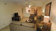 This single-floor residence showcases an open-concept floor plan that seamlessly integrates the main living spaces into one continuous, functional layout. The living room serves as the central hub, furnished with a generous sectional sofa, an armchair, and a cushioned loveseat arranged around a dark wood coffee table atop a patterned area rug. A ceiling fan with a light fixture overhead provides both comfort and illumination throughout the space. A flat-screen television sits on a low media console along one wall, adding a practical entertainment focal point.

The living area flows naturally into a dedicated dining space, where a compact dining table and chairs are positioned beneath a chandelier, accompanied by a floor lamp and wall art that adds a decorative touch. Just beyond, a pass-through counter connects to the kitchen, which features white cabinetry, a built-in microwave, and standard appliances. The kitchen's bright overhead lighting contrasts warmly with the neutral, beige tones found throughout the rest of the home. Light-colored carpeting and warm wall tones contribute to a cohesive and inviting atmosphere across the entire living area.