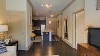 This single-floor residence showcases an open-concept design that seamlessly connects the main living areas. Dark hardwood floors run throughout, creating a rich, cohesive aesthetic that anchors the space. Crown molding adds a refined architectural detail along the ceilings, lending a polished character to every room.

The kitchen is a standout feature, equipped with dark cabinetry that extends to the upper walls, light-toned countertops, and a central island that provides additional workspace and casual seating. A pendant light hangs above the island, complementing the track lighting installed along the ceiling for layered illumination. A white refrigerator and additional appliances complete the functional kitchen setup.

The open layout extends naturally into the adjoining living and dining areas, where a patterned area rug defines the space. A decorative mirror and framed artwork are positioned along the walls, adding visual interest. Neutral-toned walls throughout keep the interiors feeling bright and airy, while multiple doorways suggest well-distributed room access. The overall ambiance is modern and inviting, balancing practical design with stylish, contemporary details.