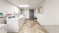 Located at 720 Queens Ave in New Westminster, BC, this shared laundry facility offers a well-organized and functional space designed to accommodate residents' laundry needs efficiently. The room is finished with attractive herringbone-patterned wood-look flooring that adds a clean, modern aesthetic throughout.

Along one wall, a row of top-loading washing machines and dryers is neatly arranged, providing ample capacity for multiple users. A commercial-grade stainless steel utility sink with a flexible spray hose is mounted near the entrance, ideal for pre-treatment or hand-washing of garments. A long stainless steel work table with a lower storage shelf runs along the opposite wall, offering generous surface space for sorting and folding laundry.

The ceiling features a drop tile grid with integrated fluorescent lighting panels that ensure bright, even illumination across the entire room. White-painted walls keep the space feeling open and clean, while a bulletin board near the entrance displays posted instructions and notices for users. A secure grey door with a lock provides controlled access to the facility. The overall layout is practical and efficient, making this laundry room a well-equipped shared amenity for the building's residents.