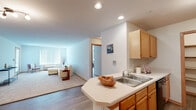 This single-floor residence offers a thoughtfully designed layout with a clean, neutral aesthetic throughout. The open-concept kitchen flows seamlessly into the living area, creating a connected and functional space. The kitchen is equipped with warm-toned oak cabinetry, a peninsula with a double-basin sink and white countertops, black appliances including a refrigerator and range, and a dedicated pantry with built-in shelving — providing ample storage. Recessed lighting illuminates the kitchen and adjacent living space, which features durable wood-look flooring.

The living area is bright and welcoming, with soft carpet, a large window fitted with vertical blinds, and a relaxed, open feel. A secondary room offers flexible use — ideal as a bedroom, home office, or fitness space — with plush carpet, a window overlooking greenery, and a baseboard heating unit. An additional carpeted room provides further versatility. The bathroom includes a wide vanity with a generous countertop, a tub-shower combination, and warm wood-trimmed accents. Throughout the space, warm wood trim, soft neutral walls, and consistent flooring choices contribute to a cohesive and comfortable living environment.