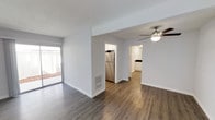 This single-floor residence offers a clean, modern aesthetic defined by an open-concept layout and consistent design choices throughout. The main living area is finished with rich, wood-style plank flooring that extends seamlessly into the adjacent spaces, creating a cohesive and inviting atmosphere. Crisp white walls and white trim provide a bright, neutral backdrop, while a ceiling fan with a built-in light fixture adds both comfort and a subtle decorative touch.

A sliding glass door in the living area allows natural light to filter in and provides direct access to an outdoor space, enhancing the connection between interior and exterior. The kitchen is efficiently positioned and features a stainless steel refrigerator, white cabinetry reaching toward the ceiling, and a clean, functional layout. A floor-level vent indicates a central air system throughout the home.

Beyond the main living space, a hallway leads to additional rooms, including what appears to be a bathroom with white cabinetry and countertops, continuing the home's consistent, contemporary style. The overall ambiance is bright, well-maintained, and move-in ready, with an emphasis on practical design and modern finishes that appeal to a wide range of occupants.