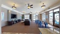 This expansive single-floor community space is thoughtfully designed to accommodate a variety of social and recreational activities. The layout is open and fluid, connecting several distinct zones while maintaining a cohesive contemporary aesthetic throughout.

The lounge areas feature plush leather seating, including tufted sofas and barrel chairs accented with bold blue throw pillows, arranged around a wall-mounted flat-screen television set into a recessed niche. Warm carpet flooring anchors these relaxation zones, while large multi-pane windows flood the space with natural light and offer views of the surrounding neighborhood.

Wood-look flooring runs through the more active areas, including a generously sized kitchen equipped with rich cherry cabinetry, granite countertops, black appliances, and a large island with bar seating. Adjacent dining areas feature high-top tables and vibrant blue metal chairs and stools, creating a casual yet stylish atmosphere.

A dedicated coffee station and small café-style seating area provide a welcoming spot for informal gatherings. Multiple ceiling fans and pendant lighting fixtures contribute to a comfortable, well-lit environment. Additional seating nooks with large windows offer quiet spaces for conversation or relaxation. Overall, the space presents a polished, functional, and inviting community environment.