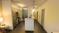 This well-appointed single-floor residence offers a thoughtfully designed open-concept layout that seamlessly connects the kitchen, dining, and living areas. The kitchen serves as the heart of the home, anchored by a central island with a granite countertop and crisp white cabinetry that extends to the ceiling. Stainless steel appliances, including a gas range, microwave, and refrigerator, add a contemporary touch, while stylish pendant lights hang above the island to create a warm, inviting atmosphere. Track lighting overhead ensures the space is well-lit throughout. Dark hardwood flooring runs the length of the unit, providing a rich contrast against the white walls and cabinetry. Crown molding adds a refined architectural detail to the ceilings. The dining area, set against a sage green accent wall, provides a cozy nook for meals, while additional rooms accessible through doorways off the main corridor offer private retreats. The overall ambiance is modern yet comfortable, with a clean, neutral palette accented by warm lighting that makes the space feel both functional and welcoming.