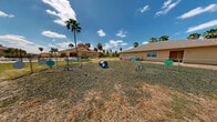 This outdoor dog park is a dedicated, fully enclosed recreational space designed for canine activity and socialization. The grounds are covered with gravel, providing a low-maintenance, durable surface suited to heavy pet use in a warm, arid climate. The park is secured by chain-link fencing that runs the full perimeter, with gated entry and exit points to ensure safe containment of dogs during use.

Throughout the space, a variety of agility and play equipment is distributed across the grounds, including tunnel crawls, hoop jumps, and weave-style obstacles, offering physical and mental stimulation for dogs of varying sizes. Colorful signage is posted at multiple points along the fencing, likely conveying park rules and guidelines for visitors.

Tall palm trees punctuate the landscape, adding a distinctive Southwest character to the environment, while the surrounding neighborhood — featuring Spanish-style architecture and residential buildings — is visible beyond the fence line. The park benefits from abundant natural light and open skies, with wide, unobstructed sightlines in all directions.

A decorative fire hydrant and waste station are also present within the space, rounding out the practical amenities. The overall layout is spacious and open, providing ample room for dogs to run freely between the installed features, making it a functional and well-equipped community gathering spot for pet owners.