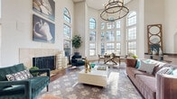 This elegantly designed single-floor space immediately impresses with its dramatic two-story ceilings and an expansive wall of floor-to-ceiling arched and gridded windows that flood the interior with abundant natural light and frame lush outdoor palm tree views. The open-concept layout seamlessly connects the living, dining, and kitchen areas, creating a cohesive and airy atmosphere throughout.

The living area is anchored by a classic tile-surround fireplace with an ornate wrought iron screen, complemented by rich hardwood flooring, a geometric patterned area rug, and a statement circular chandelier overhead. A dedicated reception or workspace nook near the entry features a built-in desk with hardwood flooring and curated wall art, offering a functional yet stylish touch.

The dining area sits beneath soaring ceilings bathed in natural light from the surrounding multi-pane windows, with views of swaying palms creating a serene backdrop. Adjacent, the kitchen is well-appointed with granite countertops, gray cabinetry, stainless steel appliances, a breakfast bar with seating, and tile flooring. An arched window above the sink adds an architectural flourish. Throughout the space, the interplay of hardwood and tile flooring, white walls, and generous natural light contributes to a bright, welcoming, and sophisticated ambiance.