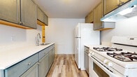 This single-floor residence offers a thoughtfully arranged layout visible in its 3D floor plan, with distinct zones for living, sleeping, and cooking. The galley-style kitchen is a functional highlight, featuring gray-toned cabinetry with brass hardware, light quartz-style countertops, a stainless steel sink, and white appliances including a gas range and full-size refrigerator. Warm wood-look laminate flooring runs the length of the kitchen, adding a modern contrast to the classic cabinetry.

The living area is bright and open, with neutral carpet flooring, white walls, and a large window that draws in natural light while offering views of the surrounding urban streetscape. Baseboard heating is installed along the perimeter, providing consistent warmth. The entry door features a classic wood finish with brass hardware.

The bedroom spaces are carpeted and neutral in tone. One bedroom benefits from dual windows and a ceiling fan, enhancing ventilation and light, with baseboard heating present as well. Another room features two side-by-side closets with louvered bifold doors, offering ample storage.

The bathroom is finished with a classic black-and-white octagonal tile floor, white subway tile surrounding the tub-shower combo, and a bold blue accent tile border, lending a timeless, clean aesthetic to the space.