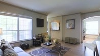 This single-floor space presents a thoughtfully designed living area that balances comfort and style through a cohesive neutral color palette. The living room serves as the focal point, featuring light hardwood flooring that extends throughout the space, complemented by soft sage-green walls that lend a calm, inviting atmosphere.

A standout architectural detail is the arched alcove set into the wall, framed by a decorative mirror that adds depth and visual interest to the room. This archway, along with crown molding accents, provides a refined touch to the overall design.

Natural light enters through a large double-hung window fitted with a roller shade, brightening the space and highlighting the warm tones of the flooring. The living area accommodates a cushioned sofa and armchair, centered around a rectangular coffee table topped with decorative accessories, all anchored by a patterned area rug in muted tones.

Additional furnishings include a compact side table used for display, and a pair of upholstered ottomans that offer flexible seating. Decorative elements such as framed wall art, potted greenery, and sculptural accents contribute to a polished, well-curated ambiance throughout.