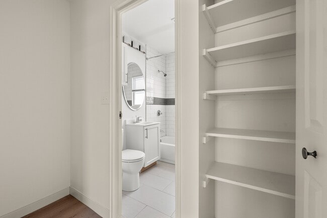 Modern bathroom with sleek white tiles, a stylish round mirror, and an adjacent open shelving unit for ample storage. - Stony Brook Commons Apartments