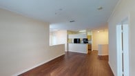 This single-floor space is defined by an open-concept layout that seamlessly connects the living area to the kitchen. Rich, dark hardwood flooring extends throughout the main living space, providing a warm contrast to the light, cream-toned walls and white textured ceiling. The generous living area offers an expansive, flexible footprint suitable for a variety of furniture arrangements.

The kitchen is positioned centrally within the layout and features a raised breakfast bar that partially encloses the cooking area while maintaining visual connectivity to the rest of the space. Warm honey-toned cabinetry lines the kitchen walls, complemented by dark granite or stone countertops and black appliances, including a refrigerator, microwave, and range. Under-cabinet lighting provides functional illumination to the workspace.

A pass-through window or opening connects an additional area to the living space, enhancing the open flow of the layout. Track lighting in the kitchen area and recessed lighting in the living space ensure consistent illumination throughout. The clean, neutral color palette and open design contribute to a bright, airy ambiance overall.
