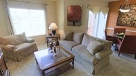 The living room serves as the heart of this single-story space, designed with comfort and classic style in mind. The room is finished with wall-to-wall carpeting in a warm neutral tone, contributing to an inviting and soft underfoot feel throughout. A plush, light gray upholstered sofa takes center stage, accompanied by a matching armchair, creating a cohesive and relaxed seating arrangement. A wooden coffee table with a glass inset panel anchors the space, while a coordinating end table holds a traditional table lamp, adding a warm ambient glow. Natural light flows in generously through multiple windows dressed with layered curtains and decorative valances, enhancing the airy feel of the room. A bold, rust-toned abstract artwork on the wall introduces a striking focal point against the neutral beige walls. Candle holders atop the coffee table add a subtle decorative touch. Access to an adjacent area is visible through a set of white paneled doors, suggesting a well-connected and functional floor plan. Overall, the space presents a traditional aesthetic with thoughtful décor details.