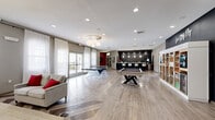 This generously sized game room spans a single floor and is thoughtfully designed to serve as a premier social and recreational hub. The open layout is anchored by light-toned hardwood-style flooring, with a striking chevron pattern in the lounge area adding a bold design element underfoot.

The space is equipped with a range of entertainment options, including a full-sized pool table, a ping-pong table, and a shuffleboard table, providing ample variety for guests. A sleek bar area features a dark accent wall, pendant lighting, and modern white bar stools, creating an inviting spot to gather. Adjacent to the bar, open shelving units display games, snacks, and beverages alongside a small refrigerator.

A dedicated lounge area is furnished with vibrant fuchsia accent chairs, a neutral sofa, and a coffee table, all centered around a large wall-mounted television set against a dramatic black tile feature wall. Neon signage reading "Live. Study. Play." reinforces the room's energetic character.

Natural light fills the space through multiple large windows dressed with sheer white curtains, offering views to the exterior. Recessed ceiling lighting and a decorative chandelier ensure the room is well-lit throughout. A glass-door entry and access to an adjacent room — appearing to be a study area — complete the layout, making this a versatile and dynamic communal space.