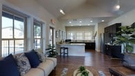 This single-story space showcases an open-concept layout that seamlessly connects the living area and kitchen, creating a cohesive and functional environment. The living area features warm hardwood flooring, vaulted ceilings that enhance the sense of openness, and large windows that allow generous natural light to fill the space. A comfortable seating arrangement anchors the living zone, complemented by decorative accents including patterned throw pillows and potted greenery.

The kitchen is a standout feature, designed with dark cabinetry contrasting against light granite countertops that extend across both the perimeter counters and a central island. The island includes bar seating, making it an ideal space for casual dining or entertaining. A stainless steel refrigerator is visible along the far wall, and multiple windows above the sink area provide additional natural light and views to the exterior. Recessed lighting throughout the kitchen ensures well-lit functionality.

The transition between the two zones is smooth and unobstructed, reinforcing the open floor plan. White walls and consistent hardwood flooring throughout create a unified, clean aesthetic, while the combination of vaulted and standard ceiling heights adds architectural interest across the space.