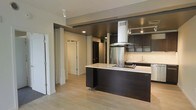 This single-floor space showcases a contemporary open-concept layout that integrates the kitchen and surrounding living areas into one cohesive environment. The kitchen serves as the centerpiece, featuring sleek dark cabinetry that contrasts sharply with light countertops on a substantial central island equipped with a built-in cooktop. A stainless steel range hood suspended from above the island adds an industrial yet refined focal point, while matching stainless steel appliances, including a full-sized refrigerator, reinforce the modern aesthetic. Upper cabinetry with under-cabinet lighting lines the walls, providing ample storage and a polished finish. Light hardwood-style flooring runs throughout, brightening the space and creating a sense of continuity between rooms. Recessed lighting is thoughtfully placed across the ceiling to ensure even illumination throughout. A hallway visible from the kitchen leads to additional rooms, hinting at a well-organized floor plan. The overall ambiance is clean, minimalist, and functional, with a neutral color palette accented by dark design elements that give the space a sophisticated, urban character.