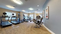This fitness center is a thoughtfully designed, single-floor facility built to support a comprehensive workout experience. The space is divided into distinct zones, each dedicated to a different aspect of physical training. The main cardio area is lined with a variety of machines, including multiple treadmills, ellipticals, and a stationary bike, many equipped with individual monitors for entertainment or performance tracking. Several windows along the walls invite natural light, creating an open and energizing atmosphere throughout.

A dedicated strength training section features a multi-station cable machine, free weights, and a weight bench, offering users ample options for resistance training. A separate curved-wall area provides additional space for functional fitness equipment, including stability balls and resistance bands, catering to stretching and accessory work. A wall-mounted flat-screen television is positioned for easy viewing from multiple points in the room.

The flooring throughout is a durable, patterned carpet in neutral tones, complementing the warm beige and soft blue wall palette. Flush-mounted circular ceiling fixtures provide consistent, even illumination across the entire space. Framed artwork adorns the walls, adding a polished, welcoming finish to the facility. The layout ensures generous open floor space, allowing users to move freely between stations without congestion.