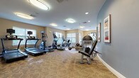 This fitness center is a thoughtfully designed, single-floor facility built to support a comprehensive workout experience. The space is divided into distinct zones, each dedicated to a different aspect of physical training. The main cardio area is lined with a variety of machines, including multiple treadmills, ellipticals, and a stationary bike, many equipped with individual monitors for entertainment or performance tracking. Several windows along the walls invite natural light, creating an open and energizing atmosphere throughout.

A dedicated strength training section features a multi-station cable machine, free weights, and a weight bench, offering users ample options for resistance training. A separate curved-wall area provides additional space for functional fitness equipment, including stability balls and resistance bands, catering to stretching and accessory work. A wall-mounted flat-screen television is positioned for easy viewing from multiple points in the room.

The flooring throughout is a durable, patterned carpet in neutral tones, complementing the warm beige and soft blue wall palette. Flush-mounted circular ceiling fixtures provide consistent, even illumination across the entire space. Framed artwork adorns the walls, adding a polished, welcoming finish to the facility. The layout ensures generous open floor space, allowing users to move freely between stations without congestion.