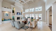 This single-story communal space showcases an open, airy design defined by dramatically soaring ceilings that rise through coffered and vaulted sections, creating an expansive, welcoming atmosphere throughout. Large transom windows and full-height glass doors line the exterior walls, flooding the interior with natural light while offering views of the surrounding outdoor amenity areas, including what appears to be a pool deck and landscaped grounds.

The entry foyer greets visitors with light neutral tile flooring, a chandelier, and a pair of accent cabinets flanking glass entry doors. This transitions seamlessly into the main lounge, where a central seating arrangement features a cream sofa, teal upholstered armchairs, and a wooden coffee table, all anchored by a vibrant striped area rug. Ceiling fans provide comfort in the grand main volume.

A prominent white fireplace with a full surround and wall-mounted television serves as a focal point along one wall, flanked by a built-in window bench seat upholstered in teal, ideal for relaxed seating. Nearby, a cozy nook features a round metallic dining table and upholstered chairs beside a built-in cushioned bench, framed by coastal artwork and decorative accents. The overall palette—soft beige, white, teal, and lime green—creates a bright, coastal-inspired ambiance throughout.