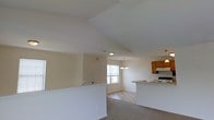 This single-floor home offers an inviting open-concept design that seamlessly connects the main living areas. The living space is distinguished by soaring vaulted ceilings that enhance the sense of openness and volume, while neutral-toned walls and light carpeting create a warm, welcoming atmosphere throughout. A half-wall partition adds subtle definition between the entry and living areas without disrupting the flow of the space. Natural light filters in through a window in the dining area, which is accented by an elegant chandelier that adds a decorative focal point overhead. The kitchen is positioned adjacent to the dining area and features warm honey-toned wood cabinetry, a light-colored countertop with a peninsula-style breakfast bar, a range with overhead ventilation hood, and tile flooring that transitions naturally from the carpeted living spaces. The open sightlines between the kitchen, dining, and living areas make this home well-suited for both everyday living and entertaining. The consistent use of soft, neutral finishes throughout lends the interior a cohesive and airy aesthetic, while the vaulted ceiling design adds architectural character and distinction to this single-level residence.