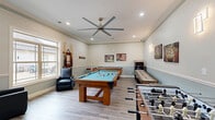 Located at 445 Warfield Blvd in Clarksville, TN, this dedicated game room is a fully equipped recreational space designed for socializing and entertainment. The room features light-toned hardwood-style flooring, two-tone walls with a soft blue wainscoting accent below a neutral upper half, and elegant crown molding that adds a polished architectural detail throughout. Recessed lighting and a large modern ceiling fan with a brushed metal finish keep the space bright and comfortable.

The centerpiece of the room is a solid wood pool table with a light blue felt surface, accompanied by a wall-mounted cue rack. Surrounding it is an impressive array of gaming options, including a foosball table, an air hockey table, and a shuffleboard table, offering entertainment for a variety of preferences. A wall-mounted flat-screen television is positioned above a low credenza with bar-height seating, creating a casual lounge area. Comfortable leather recliner chairs provide additional seating throughout.

Large windows and a glass-paneled entry door flood the space with natural light and offer views of the outdoor surroundings. Decorative artwork adorns the walls, adding personality and charm. This game room delivers a vibrant, community-focused atmosphere ideal for recreation and gathering.