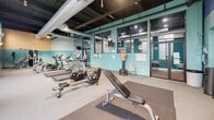 Located at 800 James Ave in Scranton, PA, this fitness center offers a generously sized, single-floor exercise room designed to support a comprehensive workout experience. The space showcases a distinctive industrial-chic aesthetic, with an exposed black ceiling revealing large ventilation ductwork and structural beams overhead, lending the room a loft-like character. The walls are painted in a bold teal hue with brick detailing, creating an energetic and motivating atmosphere.

Natural light flows in through a series of tall arched and rectangular windows along the perimeter, offering views to the outside and brightening the workout environment. The floor is finished with durable, cushioned carpeting suitable for a variety of fitness activities.

Equipment is well-distributed throughout the space and includes multiple elliptical trainers, treadmills, a rowing machine, a cable strength machine, and an adjustable workout bench. A dedicated free weights area features a full rack of dumbbells, kettlebells, and exercise mats for stretching or floor work. A heavy bag mannequin and stability balls round out the functional training options.

Wall-mounted mirrors expand the sense of space and allow for form-checking during exercise. A mounted flat-screen television and a wall clock provide additional convenience. The room is completed with recessed and track lighting, ensuring visibility throughout all hours.
