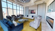 This refined lounge space exudes contemporary elegance through a thoughtfully curated blend of design elements and high-end furnishings. Soaring floor-to-ceiling windows flood the interior with abundant natural light while framing sweeping views of the surrounding urban skyline, creating a dynamic connection between the interior and the city beyond. A striking, large-scale chandelier composed of cascading metallic strands serves as a dramatic focal point overhead, anchoring the space with artistic flair. The seating arrangement is both stylish and inviting, featuring a crisp white sofa accented with navy blue throw pillows, plush gray wingback chairs, and a bold navy velvet sofa that adds a rich contrast to the overall palette. A circular metallic coffee table sits at the center, complementing the gold-toned flooring that adds warmth and depth to the space. The walls are finished with a neutral, textured treatment and framed paneling that lends a polished, sophisticated character. A wall-mounted display screen is seamlessly integrated into the design, adding a functional yet unobtrusive modern touch. Sheer floor-length drapery softens the expansive glazing and contributes to the space's refined, airy ambiance.