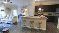 This single-floor space showcases an open-concept layout that connects the kitchen and living area in a cohesive, functional design. The kitchen is outfitted with dark espresso cabinetry, granite countertops, a tile backsplash, and stainless steel appliances, including a range and built-in microwave. A peninsula with a built-in sink extends into the living space, topped with matching granite and illuminated by a pendant light, creating a natural focal point for casual dining or entertaining. Three industrial-style bar stools line the peninsula, adding a modern yet relaxed touch. Crown molding runs along the ceiling, lending an elevated finish throughout. The hardwood-style flooring anchors the space with warmth and continuity, flowing from the kitchen into the living area. The living space is bright and inviting, benefiting from natural light through a window at the far end. A decorative cluster of round mirrors on the wall adds an artistic accent. The overall ambiance is contemporary and comfortable, balancing practical design with thoughtful aesthetic details.