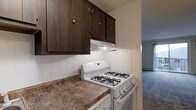 This single-floor space offers a practical and well-organized layout designed for comfortable everyday living. The kitchen serves as a central feature, equipped with rich dark wood upper cabinetry accented by brushed nickel hardware, providing ample storage. The countertops display a warm granite-style surface that complements the deep tones of the cabinetry. A white gas range with four burners sits beneath a built-in range hood, making the kitchen well-suited for cooking. The backsplash features classic white square tiles that add a clean, timeless contrast to the darker elements of the space. Under-cabinet lighting enhances visibility and adds a functional touch to the work area. A passageway visible from the kitchen leads into an adjoining area with carpeted flooring, suggesting a seamless flow between the kitchen and the living spaces beyond. The overall ambiance of the space is practical and cozy, with a layout that prioritizes functionality while maintaining a cohesive aesthetic throughout. The combination of dark cabinetry, warm countertops, and neutral tones creates an inviting environment suited to a variety of lifestyles.