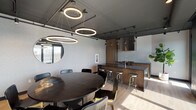 This single-floor space showcases a refined industrial-modern aesthetic, combining bold design elements with functional layouts throughout. The open-plan interior centers on a striking dining area anchored by a large, dark oval table surrounded by black-and-gold chairs. Above, a cluster of circular LED pendant lights adds dramatic visual interest, while a full chalkboard accent wall — illustrated with botanical motifs and a menu display — gives the space a creative, communal character. A round mirror reflects natural light from nearby windows, enhancing the room's depth.

The fully equipped kitchen features warm wood cabinetry paired with dark countertops, stainless steel appliances including a built-in refrigerator, gas range, wall oven, and a prominent stainless hood vent. A central island with bar-height stools provides additional seating and workspace. Light hardwood flooring runs throughout, offering a warm contrast to the predominantly dark walls and ceiling.

Floor-to-ceiling glass doors open directly onto a spacious outdoor terrace furnished with seating, offering panoramic views of the surrounding cityscape and filling the interior with abundant natural light. Track lighting and pendant fixtures provide layered illumination throughout. A connecting corridor leads to additional rooms, suggesting broader functionality within this cohesive, design-forward space.