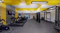 This single-floor fitness center makes a strong visual statement with its striking yellow-and-white geometric ceiling design, complemented by bright pendant lighting and durable rubber flooring throughout the main workout area. The space is thoughtfully organized into distinct zones to accommodate a variety of training styles.

The primary cardio area features a row of True-brand treadmills and elliptical machines, each equipped with individual screens for entertainment during workouts. Mounted flat-screen televisions are positioned along the walls for added convenience. The strength training section includes a multi-function cable machine, a TKO boxing bag and rack setup, a power rack, and a tiered dumbbell rack stocked with a full range of free weights.

A separate studio room, distinguished by its wood-plank flooring and exposed beam ceiling with natural light from large windows, provides a quieter, versatile space. This room houses spin bikes and a Body Solid equipment rack stocked with stability balls, medicine balls, and resistance accessories — ideal for cycling sessions, stretching, or functional fitness training.

The facility is accessed through glass-paneled double doors and features an open, spacious layout that allows for comfortable movement between equipment zones, making it suitable for a wide range of fitness levels and workout preferences.