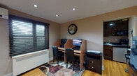 Located at 915 River Rd in New Milford, NJ, this single-floor office space is thoughtfully designed to support a productive work environment. The main work area features a dual-cubicle workstation setup with individual computer monitors, black leather chairs, and a decorative area rug over warm hardwood flooring that runs throughout the space. Recessed lighting and window blinds allow for adjustable natural light control, while a wall-mounted split-system HVAC unit ensures year-round climate comfort. A dedicated filing and printing station lines one wall, where three dark lateral filing cabinets support multiple printers, a fax machine, and additional office equipment, accented by a decorative circular wall piece above. A separate nook provides a small bistro-style table with seating for two beneath a framed floral artwork, offering a quiet spot for meetings or breaks. The compact kitchenette is well-appointed with dark wood cabinetry, granite-style countertops, a stainless steel sink, under-counter refrigerator, microwave, and a Keurig coffee maker — all the essentials for a comfortable workday. The overall ambiance is professional yet welcoming, with warm neutral tones and hardwood floors contributing to a clean, functional aesthetic throughout.
