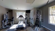 This dedicated fitness space is thoughtfully designed to support a full range of workout activities within a single-floor layout. The room features a durable checkerboard-patterned floor that adds visual interest while providing a practical, easy-to-maintain surface suited for exercise equipment. Natural light streams in through a window, brightening the space alongside recessed ceiling lighting that ensures consistent illumination throughout.

The fitness area is outfitted with a lineup of cardio equipment, including a treadmill and an elliptical trainer, positioned to take advantage of the natural light. A multi-station strength training machine anchors one side of the room, offering a variety of resistance-based exercise options. A wall-mounted flat-screen television provides entertainment or instructional content during workouts, enhancing the overall exercise experience.

The walls are finished in a neutral gray tone, contributing to a clean, focused atmosphere. A standard door provides access to an adjoining area, and the ceiling features a flush access panel alongside recessed vents, indicating a well-integrated HVAC system. The overall layout is efficient and purposeful, making this space a functional and inviting environment for daily fitness routines.