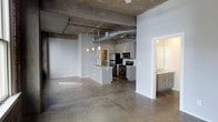 This single-floor space showcases a striking industrial-modern aesthetic that seamlessly blends raw architectural elements with contemporary finishes. The open-plan layout creates a generous, free-flowing living area anchored by polished concrete floors that reflect light and lend the space a clean, sophisticated edge.

The exposed concrete ceiling features visible structural beams, HVAC ductwork, and pendant lighting, all contributing to the loft-inspired character of the space. The kitchen is centrally positioned and designed for both function and style, featuring a peninsula island with an integrated sink, white shaker-style cabinetry, a white subway tile backsplash, and a full suite of stainless steel appliances including a refrigerator, range, and microwave.

An adjacent area — likely a bathroom or utility room — is visible through a wide doorway, outfitted with matching gray cabinetry and a countertop sink, maintaining a cohesive design language throughout. The crisp white walls throughout provide a neutral backdrop that complements the industrial ceiling elements and warm pendant lighting. The overall ambiance is modern and urban, offering a versatile canvas well-suited for a variety of uses and interior styles.