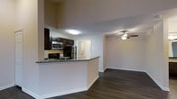 This single-floor residence offers a thoughtfully designed open-concept layout that seamlessly connects the kitchen and living areas. The kitchen is a standout feature, showcasing dark espresso cabinetry, granite countertops, and a full suite of stainless steel appliances including a gas range, microwave, and refrigerator. A breakfast bar peninsula provides both additional prep space and a natural divider between the kitchen and the adjoining living area, making it ideal for casual dining or entertaining. The living area is finished with rich, dark hardwood flooring that runs throughout, creating a warm and cohesive aesthetic. A ceiling fan overhead adds both style and practical comfort to the space. Neutral-toned walls provide a versatile backdrop, allowing for a variety of interior design preferences. A glimpse into the hallway reveals additional rooms beyond, including what appears to be a laundry or utility closet with louvered doors, suggesting practical storage and in-unit conveniences. The overall ambiance is modern and functional, with clean lines, quality finishes, and a layout designed to maximize everyday livability.