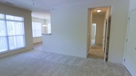 This single-floor residence features an open and functional layout with a neutral color palette of soft beige tones throughout. The main living area is finished with plush, light-colored wall-to-wall carpeting that extends into the hallway, creating a cohesive and comfortable feel underfoot. A partial wall divides the living space from the adjacent dining and kitchen area, providing a sense of definition without sacrificing the open flow between rooms. The kitchen is visible beyond the dividing wall and features a raised breakfast bar counter, ideal for casual dining or entertaining. An elegant chandelier hangs above the dining area, adding a decorative touch, while a large window allows natural light to fill the space. A carpeted hallway leads from the living area to additional rooms, with a flush-mount ceiling light providing illumination along the way. Doors visible at the end of the corridor suggest access to bedrooms or a bathroom. Throughout the space, clean white trim and smooth walls contribute to a bright, well-maintained interior with a simple, move-in-ready aesthetic.