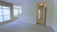 This single-floor residence features an open and functional layout with a neutral color palette of soft beige tones throughout. The main living area is finished with plush, light-colored wall-to-wall carpeting that extends into the hallway, creating a cohesive and comfortable feel underfoot. A partial wall divides the living space from the adjacent dining and kitchen area, providing a sense of definition without sacrificing the open flow between rooms. The kitchen is visible beyond the dividing wall and features a raised breakfast bar counter, ideal for casual dining or entertaining. An elegant chandelier hangs above the dining area, adding a decorative touch, while a large window allows natural light to fill the space. A carpeted hallway leads from the living area to additional rooms, with a flush-mount ceiling light providing illumination along the way. Doors visible at the end of the corridor suggest access to bedrooms or a bathroom. Throughout the space, clean white trim and smooth walls contribute to a bright, well-maintained interior with a simple, move-in-ready aesthetic.