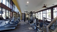 This two-story fitness center offers a comprehensive workout environment designed to accommodate a variety of exercise needs. The space is thoughtfully laid out, providing ample room for both cardio and strength training activities without feeling crowded. Large windows along one side of the facility flood the interior with natural light and offer views of the surrounding greenery, creating an inviting and energizing atmosphere.

The flooring consists of durable, patterned carpet tiles that help define different workout zones throughout the space. Cardio equipment, including ellipticals and rowing machines, is positioned along the perimeter, while the central area is dedicated to a robust selection of strength training machines and cable systems. A heavy punching bag adds a functional training option to the mix.

The ceiling features recessed lighting supplemented by a ceiling fan, ensuring consistent illumination and air circulation. Accent walls in warm yellow and deep red tones add visual character to the otherwise neutral interior. Mirrored panels along portions of the walls enhance the sense of depth and allow exercisers to monitor their form. The overall design balances functionality with a polished, modern aesthetic.