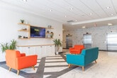 Located at 1700 Simcoe St N in Oshawa, Ontario, this single-floor lobby space offers a generously proportioned, open-concept environment that makes a bold first impression. The interior is defined by large-format marble-style porcelain tile flooring that flows throughout, complemented by a clean white drop ceiling with recessed pot lighting that ensures even, well-distributed illumination across the entire floor plate.

The space is distinguished by its vivid, energetic design language. Striking geometric murals in teal, yellow, orange, and grey adorn multiple accent walls, creating dynamic focal points at both the main entrance and along the corridors. A prominent branded orange wall near the glazed entry doors reinforces the building's identity as "17 Hundred Simcoe North."

A comfortable lounge area anchors the central zone, furnished with retro-styled red armchairs and a teal sofa arranged around a patterned area rug. Adjacent to this is a meeting or dining nook featuring a white table, modern blue chairs, and a bold yellow geometric accent wall, lit by a contemporary multi-arm pendant fixture. Built-in cabinetry with open wood shelving, integrated display screens mounted on teal accent walls, and lush tropical plants add warmth and functionality throughout. Elevator banks set against a marble-clad wall provide vertical access, while large glazed storefronts allow natural light to penetrate deep into the lobby.