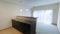 This single-floor space showcases a thoughtfully designed open-concept layout that seamlessly connects the kitchen and living area. The kitchen is outfitted with dark cabinetry that contrasts cleanly against light-colored walls, creating a modern, contemporary aesthetic. A continuous dark countertop runs the length of the kitchen, incorporating a stainless steel sink and integrated dishwasher. The countertop extends to form a peninsula-style divider, naturally separating the kitchen from the adjacent living space while maintaining an open, airy feel. Two globe-style pendant lights hang above the peninsula, providing focused task lighting and adding a stylish focal point to the space. The living area transitions smoothly from the kitchen, featuring carpeted flooring that offers a comfortable contrast to the hard flooring in the kitchen zone. A large window fitted with vertical blinds dominates the far wall of the living area, flooding the space with natural light and providing access or views to the exterior. The high ceilings throughout contribute to a spacious, open atmosphere, enhancing the overall sense of volume and airiness across both areas.