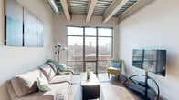 Located at 601 R St in Lincoln, Nebraska, this modern 3-bedroom, 2-bathroom unit showcases an industrial-chic aesthetic with a thoughtfully designed single-floor layout. The living room is a standout space, anchored by dramatic floor-to-ceiling windows that flood the room with natural light and frame sweeping views of Lincoln's urban skyline. Exposed metal ceiling beams and corrugated steel panels add an architectural edge, complemented by warm hardwood-style flooring throughout. The open-concept kitchen is well-appointed with light maple cabinetry, stainless steel appliances including a refrigerator, range, microwave, and dishwasher, and generous counter space. A central island with seating offers a natural gathering point and flows seamlessly into the dining and living areas, enhanced by pendant lighting overhead. The bedrooms are bright and efficiently designed, each with windows that invite natural light and closet storage with built-in shelving and hanging rods. The bathrooms feature clean, modern finishes with light wood vanities and wide mirrors. A connecting hallway ties the layout together with ease. This unit offers a compact yet functional urban living experience in the vibrant Haymarket neighborhood of Lincoln.