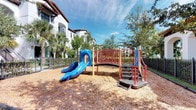 This outdoor community space centers around a vibrant playground designed for younger residents and visitors. The play structure features a prominent blue double slide alongside a raised platform accessible via steps with red safety railings, offering an engaging environment for children. The ground is covered with a generous layer of wood chip mulch, providing a safe, impact-absorbing surface throughout the play area.

The space is enclosed by a sturdy dark metal fence, ensuring a secure environment. Mature tropical trees, including palm varieties, border the perimeter, offering natural shade and a lush, verdant backdrop. The surrounding architecture is characterized by a Mediterranean-inspired aesthetic, with white stucco exteriors, red tile rooflines, and balconied facades visible in the background, lending the area an upscale residential ambiance.

The open-sky setting allows for ample natural light throughout the day, making the playground bright and inviting. The combination of the colorful equipment, natural wood chip surfacing, tropical greenery, and well-maintained surroundings creates a welcoming and safe recreational space well-suited for active outdoor play within the community.