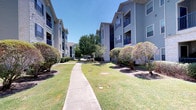 This residential community presents a welcoming exterior characterized by multi-story buildings finished in a cool gray-blue siding with contrasting dark trim accents. The buildings are arranged to create an open corridor between them, fostering a sense of community while maintaining privacy. Private balconies extend from the upper levels, offering residents outdoor space with views of the surrounding grounds.

The landscaping is a notable feature of this property, with well-manicured green lawns lining both sides of a central concrete walkway that connects the buildings. Mature trees and neatly trimmed shrubs are thoughtfully distributed throughout the grounds, providing natural shade and visual interest. The greenery creates a calm, park-like atmosphere throughout the common areas.

The walkway itself is clean and clearly maintained, providing easy pedestrian access through the property. Exterior stairwells and covered entry points are visible on the buildings, contributing to the functional layout of the community. Under bright, clear skies, the overall ambiance of the space is open, airy, and well-kept, reflecting attentive property maintenance.