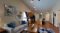 This single-floor residence showcases an open-concept design that seamlessly connects the living, dining, and kitchen areas into one cohesive and functional space. The living room draws immediate attention with its dramatically vaulted ceilings, which amplify the sense of space and airiness throughout. A ceiling fan is mounted at the apex of the vault, complementing the recessed lighting that provides warm, even illumination. The living area is anchored by a comfortable gray sofa accented with teal throw pillows, a wooden coffee table, and a patterned area rug, all set against rich hardwood flooring that extends throughout the main living space. Framed artwork on the walls adds a personal, decorative touch. The kitchen features warm-toned cabinetry, black appliances including a range and built-in microwave, and a dark countertop, with a window that invites natural light. A kitchen island with a light granite surface serves as a natural transition into the dining area, where a pendant light hangs above a dining table surrounded by dark wooden chairs. The overall ambiance is warm, modern, and inviting, with a thoughtful layout that promotes both comfortable daily living and easy entertaining.
