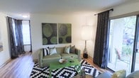 This single-floor space showcases a well-appointed living area designed with a distinct, cohesive aesthetic. The room centers around a sage green sofa adorned with an assortment of decorative pillows, including black-and-white striped and patterned designs that complement the overall color scheme. A bold black-and-white chevron area rug anchors the seating arrangement, adding graphic contrast to the warm hardwood-style flooring beneath.

A matching green coffee table serves as a functional focal point, styled with decorative objects that reinforce the room's curated look. Abstract wall art in tones of green, black, and olive adds visual depth to the neutral walls, while a tall floor lamp with a classic drum shade provides warm ambient lighting. Dark patterned drapes frame what appears to be a window, adding texture and a sense of enclosure to the space.

The textured ceiling adds a traditional architectural detail, and the overall layout feels intentional and compact yet comfortable. The consistent use of green, black, and white throughout the furnishings, art, and accessories creates a unified, design-forward environment with a modern yet approachable ambiance.