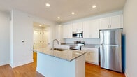 Located at 5151 Ebright Rd in Canal Winchester, Ohio, this well-appointed one-bedroom, one-bathroom unit offers a thoughtfully designed single-floor layout. The kitchen serves as a central highlight, featuring white shaker-style cabinetry extending to the ceiling, granite countertops, a spacious center island with a built-in sink, and a full suite of stainless steel appliances including a range, microwave, and refrigerator. Recessed lighting illuminates the space, complementing the warm hardwood-style flooring that runs throughout the main living areas. The living room is bright and welcoming, with a glass-paneled exterior door topped by a transom window and an adjacent window that invite natural light and provide outdoor views. The bedroom offers a comfortable, carpeted retreat with a large window overlooking the surrounding neighborhood and a clean, panel-style door leading to the closet. The bathroom is finished with a granite-topped vanity, wood-toned flooring, and a walk-in tiled shower. A hallway connects the rooms efficiently, maintaining a clean and organized flow throughout the unit. The overall aesthetic is modern and neutral, with a consistent use of white trim, soft gray walls, and quality finishes throughout.