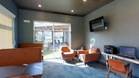 This single-story space functions as a comfortable communal lounge or leasing office area, thoughtfully designed to balance functionality with a welcoming atmosphere. The room is finished with neutral gray walls and recessed ceiling lighting that provides consistent, even illumination throughout the space. Patterned carpet in cool blue-gray tones anchors the seating area, adding a subtle decorative element underfoot.

A prominent feature of the room is the large sliding glass door, which floods the interior with natural light and offers a direct view of an outdoor patio. The patio, visible through the glass, is furnished with additional seating beneath a covered pergola structure, extending the usable space outdoors. This connection between interior and exterior enhances the overall sense of openness.

The seating arrangement centers on two mid-century modern-style armchairs in warm orange upholstery, paired with a small side table — creating an inviting conversation area. A wall-mounted flat-screen television is positioned prominently on the adjacent wall, adding a practical amenity to the lounge. Along the perimeter, a workstation with a printer suggests a functional, multi-purpose use for the space. Overall, the room strikes a balance between comfort and utility.