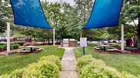 Located in Annapolis, Maryland, this outdoor Grill Station is a well-appointed communal space designed for outdoor dining and gathering. At its centerpiece stands a built-in grilling station constructed with a handsome stacked-stone base, paired with a stainless steel grill, offering a durable and attractive setup for cooking outdoors. A concrete walkway leads directly to the grill area, flanked by neatly maintained garden beds filled with flowering shrubs and ornamental plantings that add color and texture to the space. Overhead, large blue shade sail canopies are stretched between white posts, providing generous coverage from the sun and creating a comfortable, shaded environment for outdoor use. Multiple picnic tables are positioned throughout the area, offering ample seating for groups. The surrounding grounds are expansive and well-kept, featuring manicured green lawns, mature trees, and mulched planting beds that contribute to a clean, park-like atmosphere. Iron fencing defines the perimeter, while brick residential buildings frame the backdrop of the space. The overall ambiance is welcoming and functional, offering a thoughtfully landscaped outdoor retreat well-suited for community gatherings and al fresco dining.