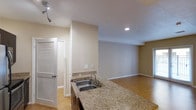 Located at 1670 Kettner Blvd, Unit #124 in San Diego, this single-floor residence offers a thoughtfully designed open-concept layout that seamlessly connects the kitchen and living areas. The kitchen is appointed with granite countertops featuring warm earth-tone veining, a double stainless steel sink, and dark cabinetry, providing both functionality and visual appeal. A peninsula counter extends the workspace and creates a natural transition into the open living area, making it ideal for entertaining or everyday use. The living space is finished with warm cork flooring, which adds a natural texture underfoot while contributing to the inviting ambiance of the home. Neutral beige and gray wall tones throughout provide a clean, versatile backdrop. Recessed lighting and a track light fixture ensure well-distributed illumination across the space. A louvered bifold door is visible near the entryway, providing access to a closet for added storage convenience. The overall design is clean and contemporary, with a cohesive material palette that balances practicality with comfort, making this San Diego unit a well-appointed and welcoming living space.