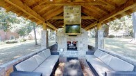 Located at 804 W Morgan St in Raleigh, NC, this outdoor pavilion is a standout amenity designed for relaxation and entertainment. The structure is anchored by a dramatic floor-to-ceiling stone fireplace that serves as the visual centerpiece of the space, with a wall-mounted flat-screen TV positioned just above it for an exceptional outdoor viewing experience. The pavilion's soaring vaulted ceiling is crafted from warm, natural wood beams and timber framing, creating an inviting, lodge-like atmosphere while providing ample shelter. Stone columns at each corner lend the structure a solid, architectural presence that complements the surrounding landscape. The flooring features classic brick pavers, adding a timeless, rustic character underfoot. Generous wicker sectional sofas with plush cushions are arranged around a sleek coffee table, offering comfortable seating for a large group. The open-air sides of the pavilion allow for natural breezes and unobstructed views of the lush, tree-lined grounds beyond. Whether used for social gatherings or quiet evenings by the fire, this pavilion delivers a refined yet relaxed outdoor living experience in the heart of Raleigh.